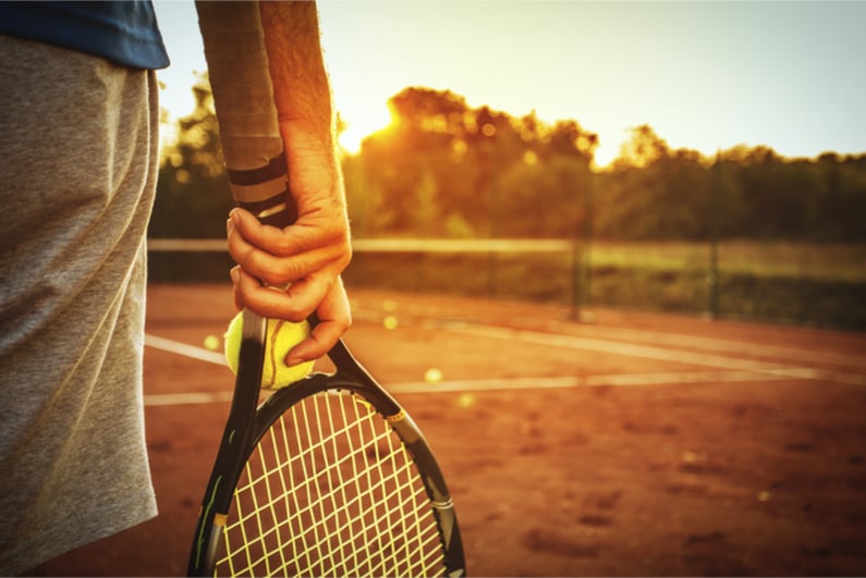 Hand holding tennis ball and racquet on clay court background